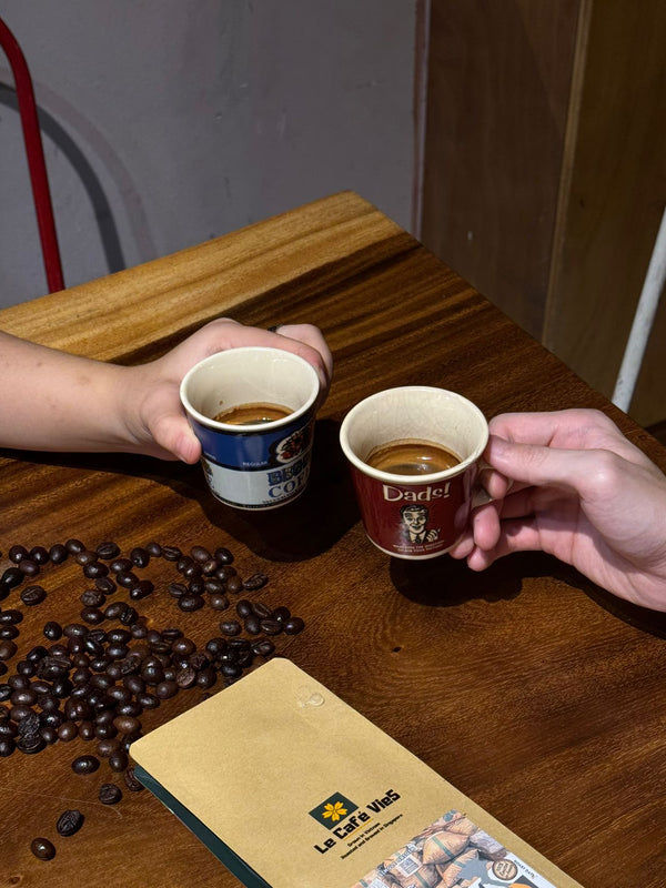 Two hands holding coffee cups with a package of coffee beans on a wooden table.