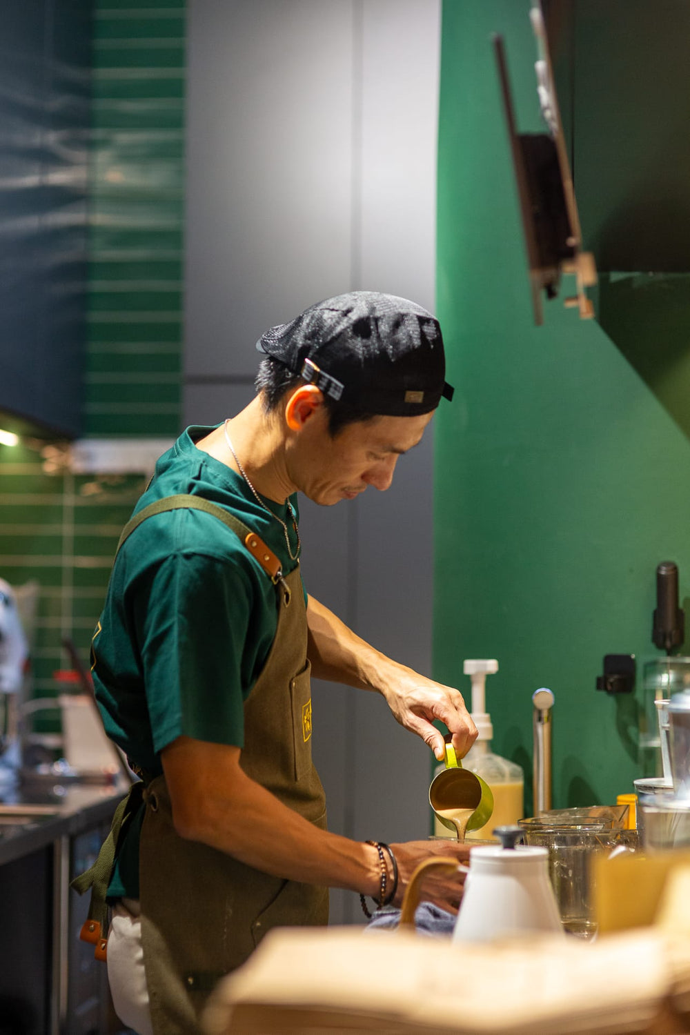 Barista in a green shirt and brown apron preparing a drink in a coffee shop.